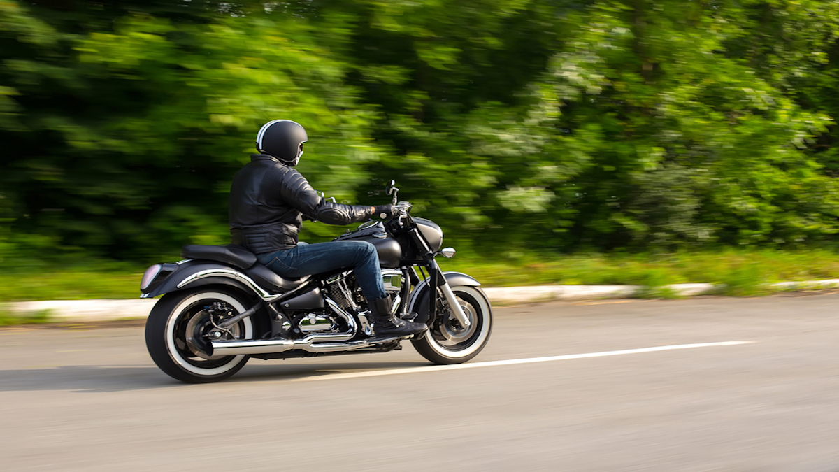 A motorcyclist drives along a road lined with trees. He's wearing a helmet, leather jacket, and dark jeans.